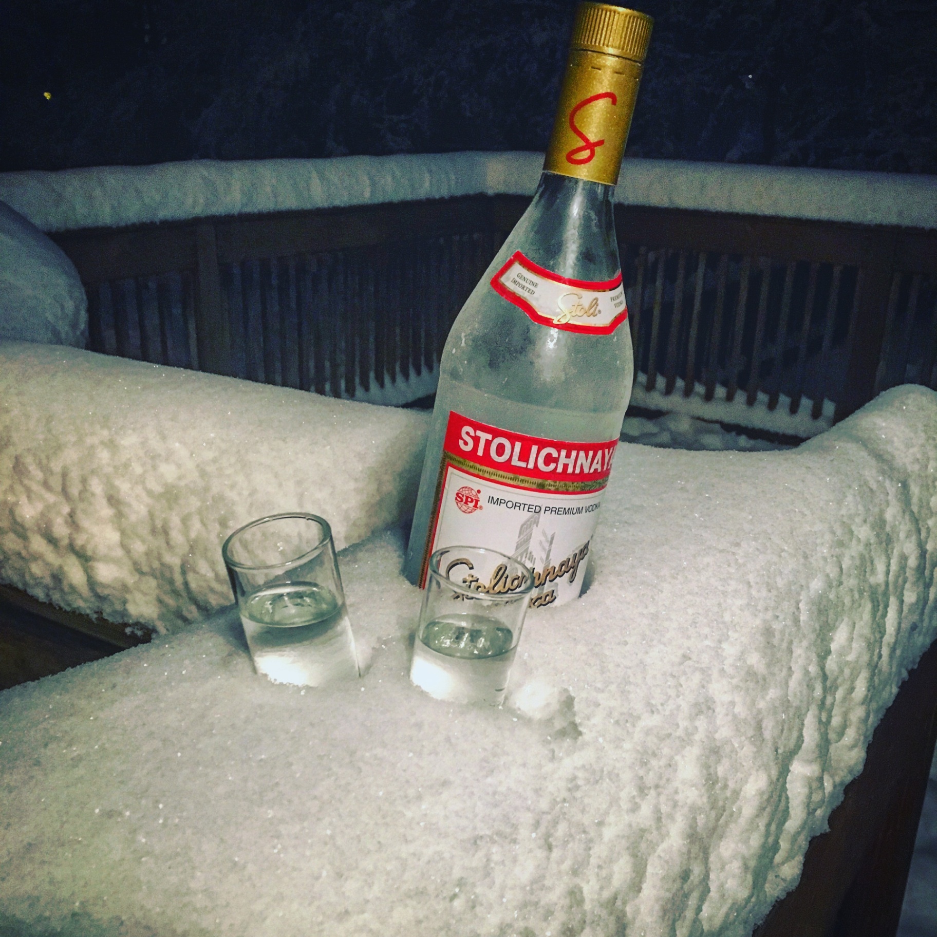 Product photography of a Stolichnaya vodka bottle and two shot glasses chilled in fresh snow at night.